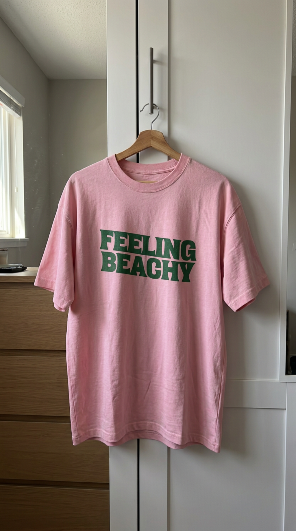 Pink t-shirt with 'FEELING BEACHY' text on a hanger against a white wall.