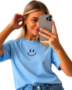 Woman wearing a light blue t-shirt with a smiley face design, taking a selfie.