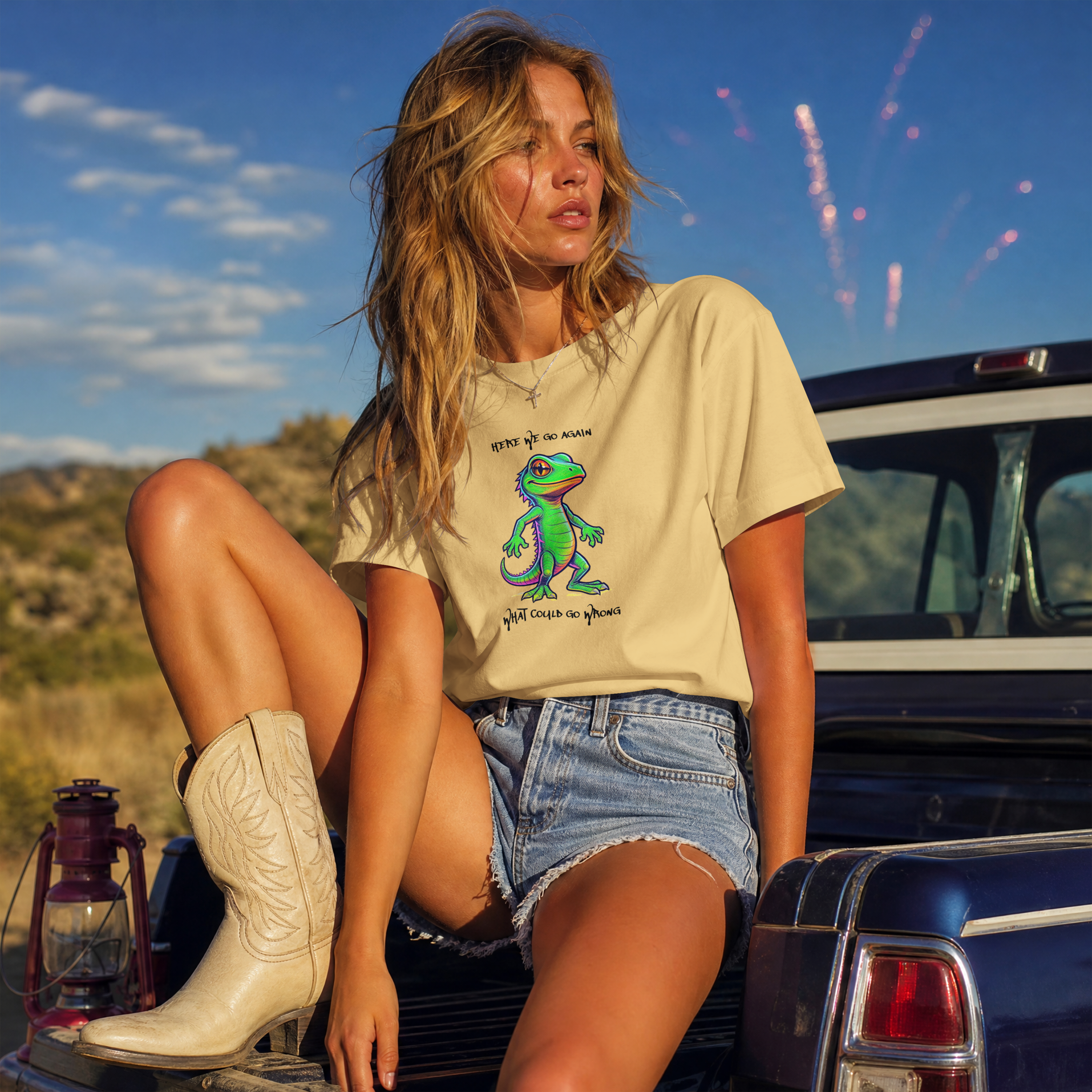 A woman sits on the tailgate of a blue pickup truck outdoors under a blue sky with fireworks. She is wearing a ONVR beige graphic t-shirt with a green gecko design and the text 'give it to me she'll love it too', denim shorts, and white cowboy boots.
