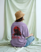 A person sitting on a draped mint green fabric set, viewed from behind. They are wearing a lavender graphic ONVR t-shirt with a large red and black 'Born to be' design on the back, paired with a tan bucket hat and blue jeans.
