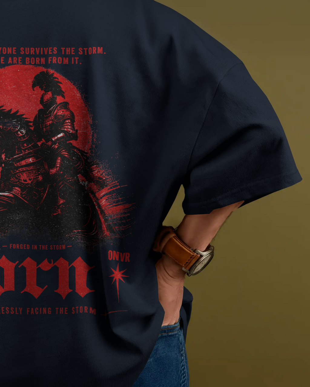 A close-up of a person wearing a navy blue graphic ONVR Oversized t-shirt with a large red and black print on the back, paired with blue jeans and a brown leather watch strap, set against a solid olive green background.