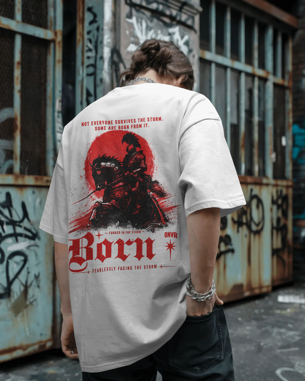 A person viewed from behind wearing a light gray ONVR Fashion oversized T-shirt with a large red and black graphic print on the back, standing in front of a weathered graffiti-covered metal wall.