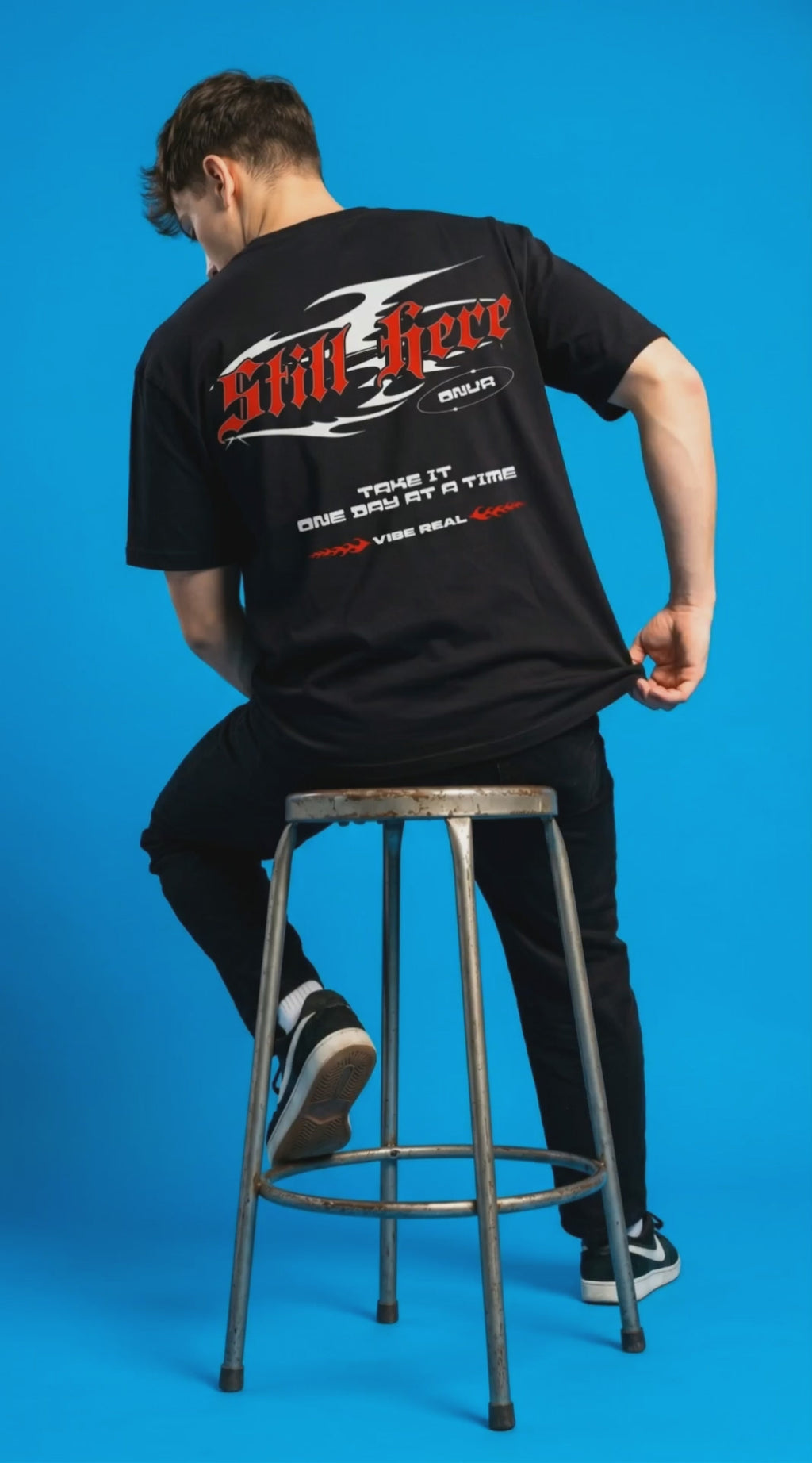 Person wearing a black t-shirt with text on the back, sitting on a stool against a blue background