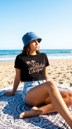 Person sitting on a beach wearing a black t-shirt with 'Retro Escape' text and a blue bucket hat.