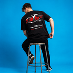 Person wearing a black t-shirt with text on the back, sitting on a stool against a blue background